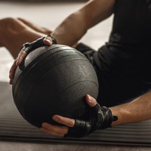 Crop unrecognizable fit male athlete in hand wraps doing twists with weighted ball on mat in gymnasium